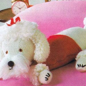 May include: White knitted poodle toy with a red knitted vest, lying on a pink blanket. The poodle has a red bow on its head and black eyes.