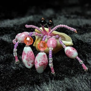 Może przedstawiać: Ręcznie robiona figurka kraba pustelnika z koralików. Krab ma różowe i fioletowe koraliki na ciele i nogach, z pomarańczowymi i czarnymi akcentami. Jest umieszczony na naturalnej muszli.