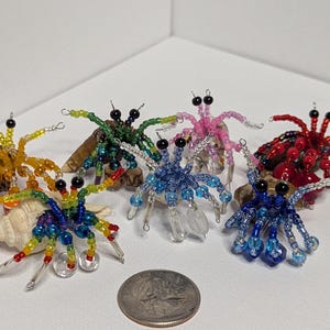 Może przedstawiać: Kolekcja koralikowych figurek krabów pustelników w różnych kolorach, w tym żółtym, zielonym, różowym, niebieskim i czerwonym. Każdy krab ma czarne oczy i jest umieszczony na małej muszli lub kamieniu. Moneta służy jako odniesienie do rozmiaru.