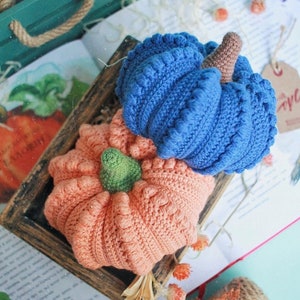 May include: Two crocheted pumpkins, one blue and one peach, sitting on a book with a wooden frame around them.