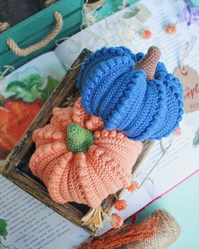 May include: Two crocheted pumpkins, one blue and one peach, sitting on a book with a wooden frame around them.