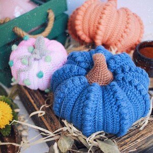 May include: Three crocheted pumpkins in different colors: pink with green accents, orange, and blue. The pumpkins are sitting on a wooden surface with straw and leaves.