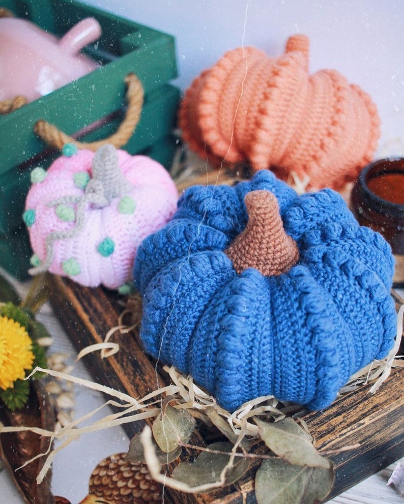 May include: Three crocheted pumpkins in different colors: pink with green accents, orange, and blue. The pumpkins are sitting on a wooden surface with straw and leaves.
