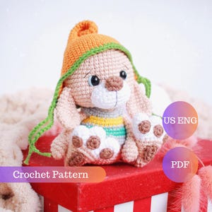 May include: Crochet pattern for a cute amigurumi bunny wearing an orange and green hat. The bunny is sitting on a red box with white stripes. The pattern is available in US English and PDF format.