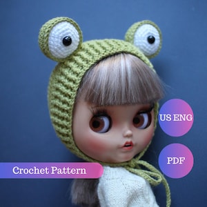 May include: A doll wearing a green crocheted frog hat with large white and black eyes. The hat is tied under the chin with a green string. The text "Crochet Pattern" is visible on a pink and purple banner.
