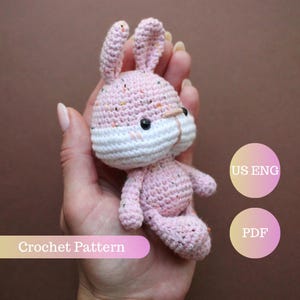 Può includere: Un coniglietto rosa all'uncinetto con muso bianco e dettagli maculati. Il coniglietto ha occhi neri ed è seduto. L'immagine include il testo "Crochet Pattern", "US ENG" e "PDF".