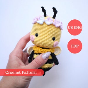 May include: Crochet pattern for a cute bee amigurumi toy. The bee is yellow and black with a pink and white flower crown. The pattern is available in US English and PDF format.