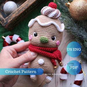 May include: Crocheted gingerbread person amigurumi toy with a red scarf and white icing. The toy is holding a bell in each hand. The toy is sitting on a wooden surface with a candy cane and a gold ornament in the background. The text "Crochet Pattern" and "Olya Povzun" are visible in the image. The text "US ENG" and "PDF" are also visible in the image.