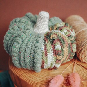 May include: A crocheted pumpkin with a white stem, green, brown, and white stripes, and a speckled green texture. The pumpkin is sitting on a wooden surface with pink dried flowers.