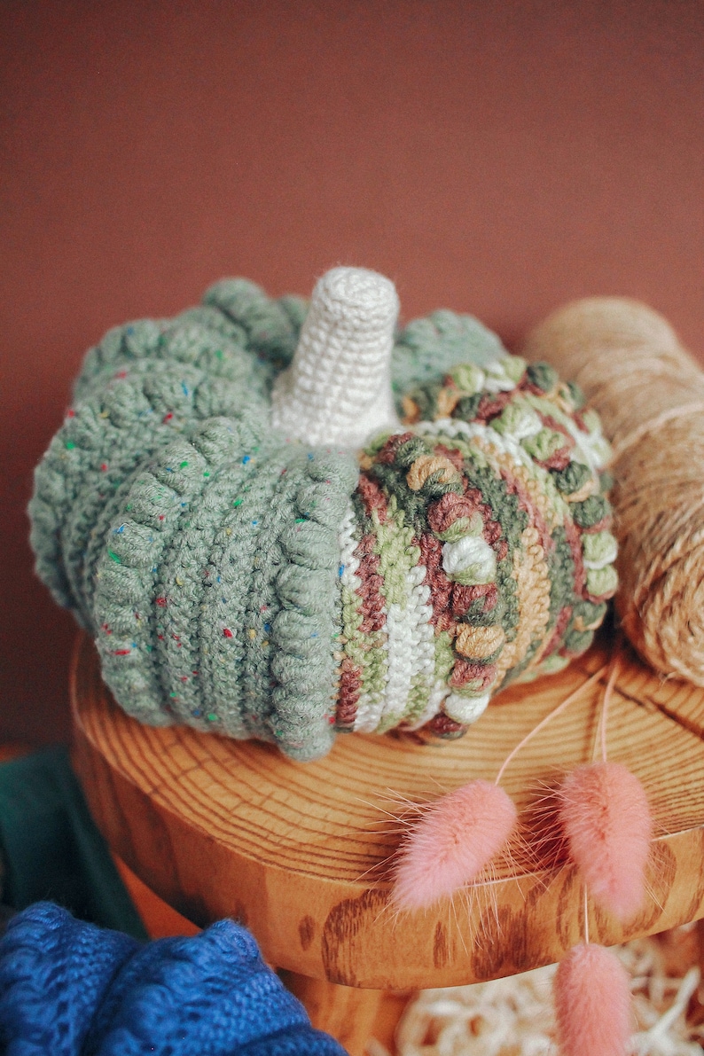 May include: A crocheted pumpkin with a white stem, green, brown, and white stripes, and a speckled green texture. The pumpkin is sitting on a wooden surface with pink dried flowers.