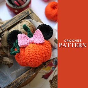 Puede incluir: Una calabaza naranja de ganchillo con orejas de ratón negras y un lazo rosa. La calabaza es parte de un patrón de ganchillo. El texto "CROCHET PATTERN" se muestra en blanco sobre un fondo naranja.