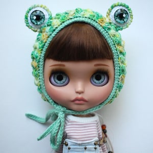 May include: A doll with brown hair and blue eyes wearing a green and yellow variegated crochet bonnet featuring two protruding frog eyes on top. The bonnet has a light green crocheted trim and ties under the chin. The doll wears a striped shirt.
