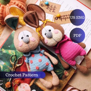 Puede incluir: Dos muñecos de conejito de ganchillo hechos a mano con detalles en marrón, beige, rosa y azul. Uno lleva un vestido azul con detalles de flores rosas y el otro un conjunto rosa. La imagen también incluye una calabaza, tijeras y etiquetas.