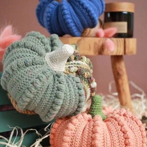 May include: Three crocheted pumpkins in different colors and patterns. The pumpkins are blue, green, and peach. They are sitting on a wooden surface.