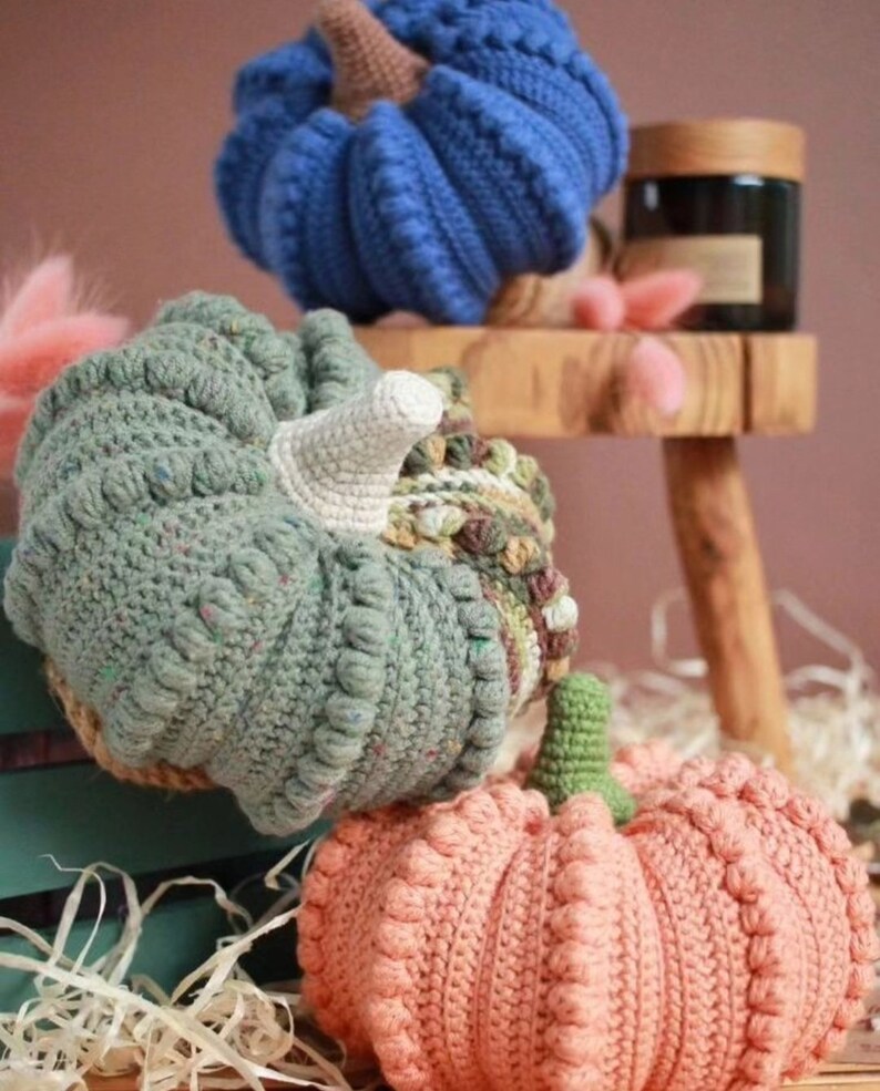 May include: Three crocheted pumpkins in different colors and patterns. The pumpkins are blue, green, and peach. They are sitting on a wooden surface.