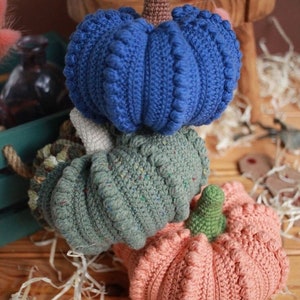 May include: Three crocheted pumpkins stacked on top of each other. The top pumpkin is blue, the middle pumpkin is green, and the bottom pumpkin is peach.