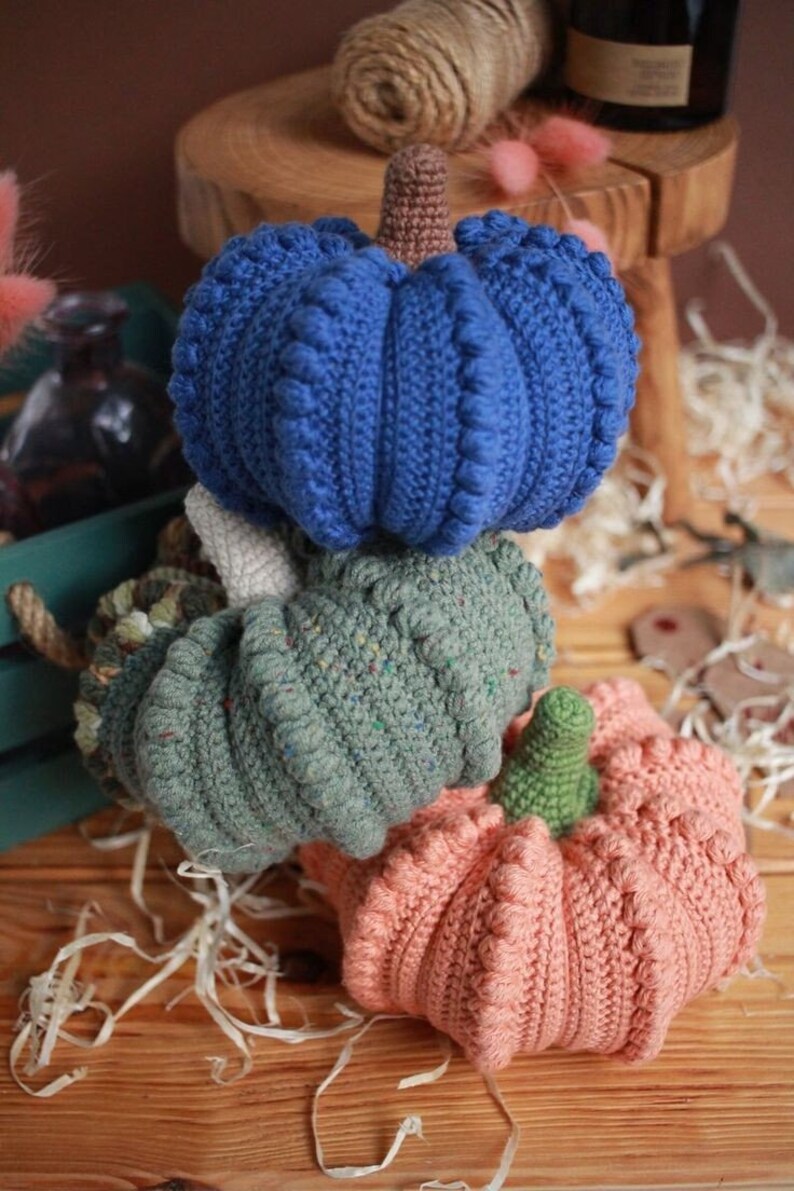 May include: Three crocheted pumpkins stacked on top of each other. The top pumpkin is blue, the middle pumpkin is green, and the bottom pumpkin is peach.