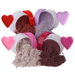 May include: Four white containers with red, pink, and purple lids are tipped over, spilling out colourful powders. The powders are in shades of burgundy, lavender, and light pink. Four heart-shaped decorations in red, pink, and purple are also present.