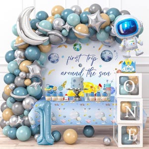 May include: A blue and silver balloon arch with stars and planets, a cake with a blue frosting, and a backdrop with the text "first trip around the sun". There are also clear boxes with the letters "O N E" and a silver astronaut balloon.