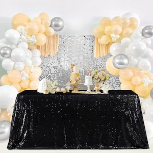 May include: A black sequin tablecloth with a white and cream balloon arch backdrop. The balloon arch is decorated with silver balloons. There are two cakes on a table in front of the tablecloth.