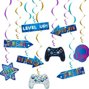 May include: A collection of gaming-themed party decorations, including blue and white controller cutouts, star-shaped bonus signs, and arrow-shaped signs with phrases like "Game On" and "Go Win." The decorations are suspended with colorful streamers.