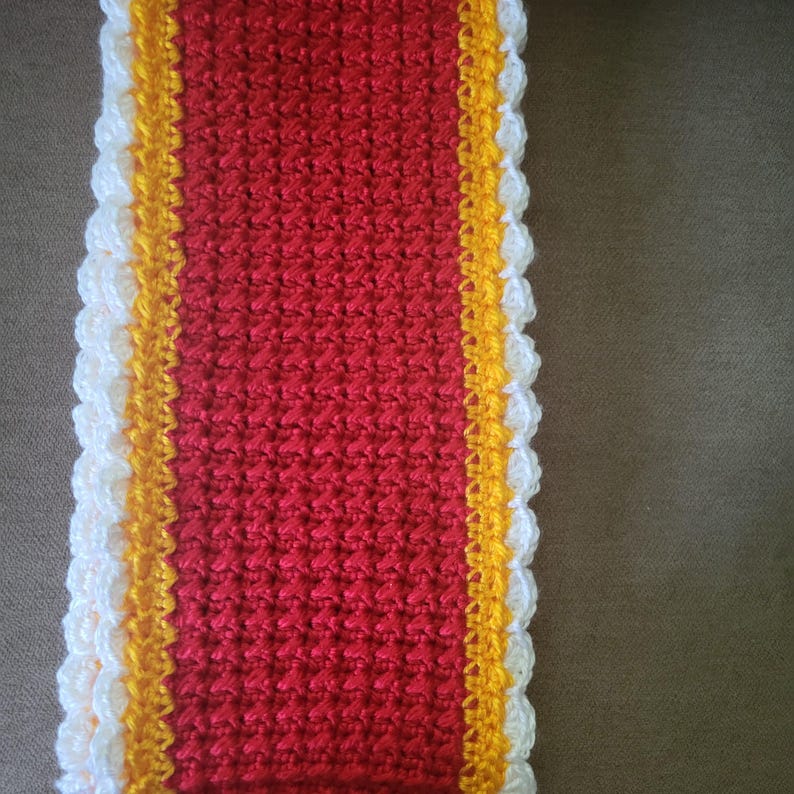 May include: A red crocheted scarf with a white and yellow border. The scarf is made with a textured stitch.