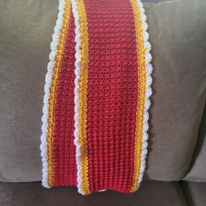 May include: A red crocheted scarf with white and yellow trim. The scarf is folded in half and lying on a brown couch.