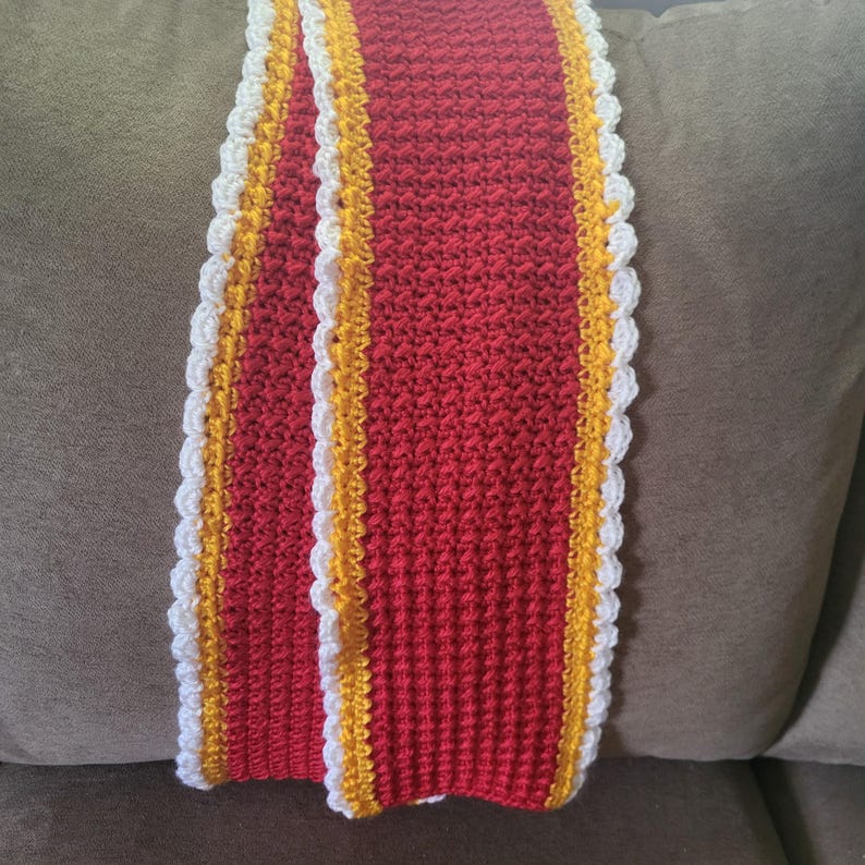 May include: A red crocheted scarf with white and yellow trim. The scarf is folded in half and lying on a brown couch.