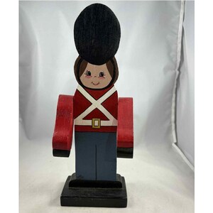 Puede incluir: Figura de soldado de juguete de madera pintada. Presenta un sombrero negro, una chaqueta roja y pantalones azules. El soldado tiene una cara pintada y un diseño de correas cruzadas blancas. La figura se apoya sobre una base negra.