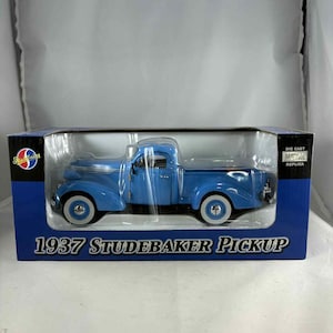 Könnte beinhalten: Ein Druckguss-Metallnachbau eines Studebaker Pickup von 1937. Das Modell ist in leuchtendem Blau mit Weißwandreifen lackiert und wird in einer blauen und transparenten Kunststoffbox präsentiert. Der Text "1937 Studebaker Pickup" ist auf der Box aufgedruckt.