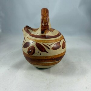 May include: A small, round, ceramic pitcher with a handle and spout. The pitcher is painted with a cream-colored base and decorated with brown horizontal stripes and leaf-like patterns. The handle is a darker brown.