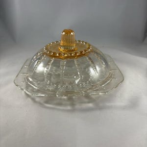 以下が含まれることがあります： フリルのあるベースと溝のある蓋が付いた透明なガラス製のバターディッシュ。蓋には小さな琥珀色のノブが付いています。