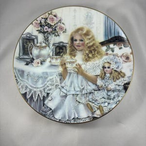May include: A porcelain plate featuring a painted image of two girls in white dresses enjoying a tea party. The girl on the left is holding a teacup and saucer. The girl on the right is holding a doll. There are pink roses in a vase behind them.