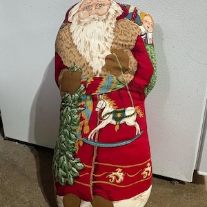 May include: A red and white Santa Claus cushion featuring a rocking horse, a Christmas tree, and a doll. The cushion is made of fabric and has a festive design.