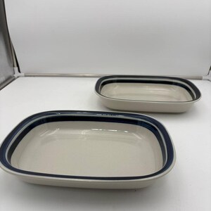 May include: Two rectangular ceramic serving dishes. The dishes are off-white with a navy blue stripe around the rim. The dishes are empty and sit on a white surface.
