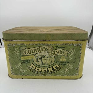 Op de afbeelding: Rechthoekige metalen broodtrommel met een groen en geel ontwerp. De trommel heeft de woorden "Country Style Bread" en "Finest Quality" in een decoratieve banner. De trommel is gemarkeerd "L.T. Putman & Sons, Est. 1880."
