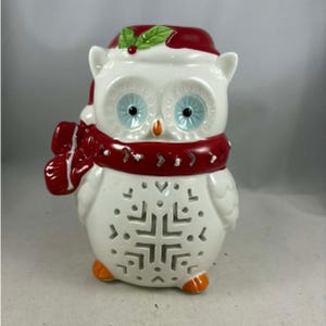 Puede incluir: Figura de búho de cerámica con un diseño festivo. El búho es blanco con ojos azules, pico naranja y pies naranjas. Lleva una bufanda roja y un gorro de Papá Noel con acebo. El pecho del búho presenta un diseño de copo de nieve.