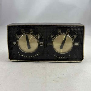 Puede incluir: Un medidor de temperatura y humedad negro vintage. El medidor tiene dos esferas circulares con centros dorados y números negros. La esfera izquierda indica "TEMPERATURE" y la derecha "HUMIDITY".