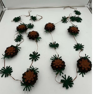 Puede incluir: Dos guirnaldas decorativas con piñas de fieltro marrón y hojas de fieltro verde. Las piñas están unidas a una fina cuerda marrón, con hojas verdes intercaladas. Ideal para la decoración navideña o de temporada.