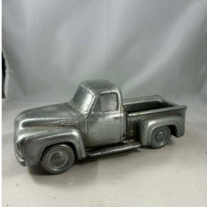 Puede incluir: Figurita de camioneta de estilo vintage de color plateado. La camioneta presenta un diseño clásico con cabina, caja y ruedas detalladas. El acabado metálico le da un aspecto rústico y antiguo. Es un objeto decorativo.