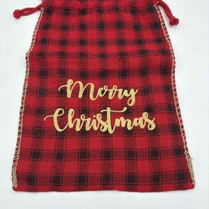 May include: Red and black buffalo plaid drawstring gift bag with the words "Merry Christmas" in gold glitter script. The bag has a burlap trim and red drawstring closure. A festive holiday-themed gift bag.