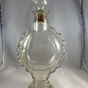 Puede incluir: Una botella de vidrio transparente con un diseño festoneado y un tapón decorativo. La botella está vacía y tiene un corcho en el cuello.