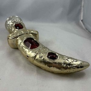 May include: A decorative, gold-colored bottle stopper with a curved design. It features three inset, rectangular, red-colored gemstones. The stopper has ornate, swirling patterns and a silver-colored top.