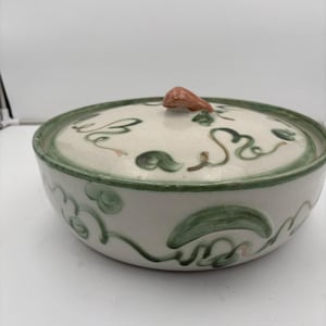 May include: A white ceramic serving bowl with a lid. The bowl has a green rim and is decorated with green vine-like patterns. The lid features a brown handle. The bowl is suitable for serving food.