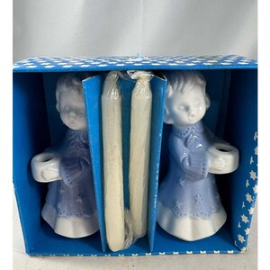 Può includere: Un set in scatola di due portacandele a forma di angelo in ceramica blu e bianca. Gli angeli indossano vesti blu con dettagli a stella e tengono in mano piccoli portacandele bianchi. La scatola è blu con un motivo a stelle bianche.