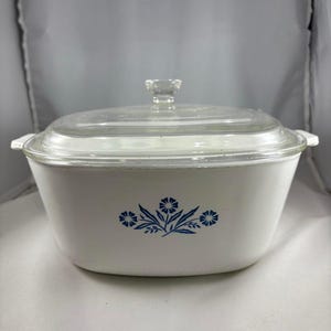 May include: White casserole dish with a clear glass lid and handle. The dish features a blue floral design on the front. The lid has a clear glass knob. The dish is designed for cooking and serving.