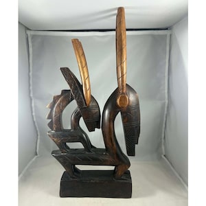 Puede incluir: Una escultura de madera marrón oscuro que presenta dos figuras estilizadas con cuellos y cuernos alargados. La escultura tiene intrincados detalles tallados y se apoya sobre una base rectangular. Los cuernos son de un tono marrón más claro.