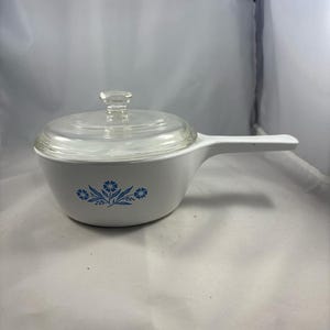 Könnte beinhalten: Ein weißer Keramiktopf mit einem Glasdeckel und einem blauen Blumendesign. Der Topf hat einen Griff an der Seite.