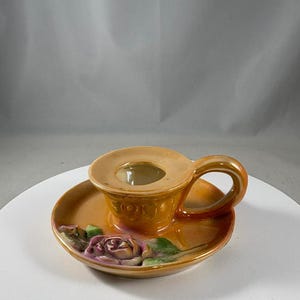 Pode incluir: Um castiçal vintage em tom laranja quente, com um topo em forma de xícara e uma alça, colocado sobre um pires. O pires é decorado com um desenho de rosa pintada em rosa e verde.