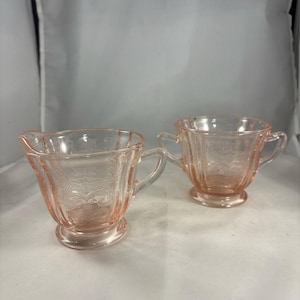 May include: A pink glass creamer and sugar bowl set. The creamer has a spout and handle, while the sugar bowl has two handles. Both pieces feature a decorative etched pattern and sit on a round base. The set is made of transparent pink glass.